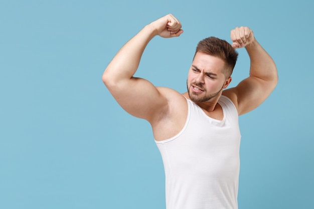 Strong young bearded guy 20s in white singlet posing isolated on pastel blue wall background studio portrait. Sport fitness healthy lifestyle concept. Mock up copy space. Showing biceps, muscles.