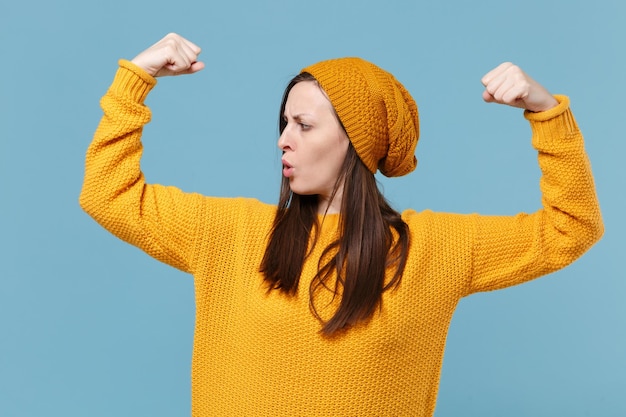 Photo strong young brunette woman girl in yellow sweater and hat posing isolated on blue background studio portrait. people sincere emotions lifestyle concept. mock up copy space. showing biceps, muscles.