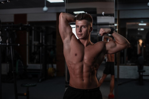 Strong young man bodybuilder with a hairstyle and a bare muscular torso trains and poses in the gym against a dark background