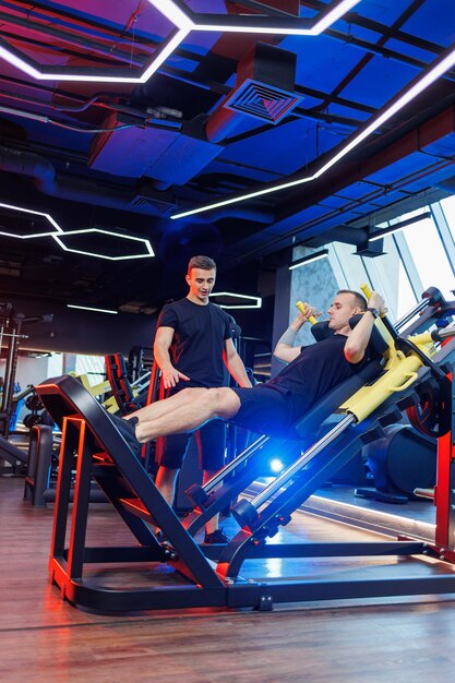 A strong young man performs exercises in the gym under the supervision of a trainer. Powerful workout by a handsome man. Healthy lifestyle