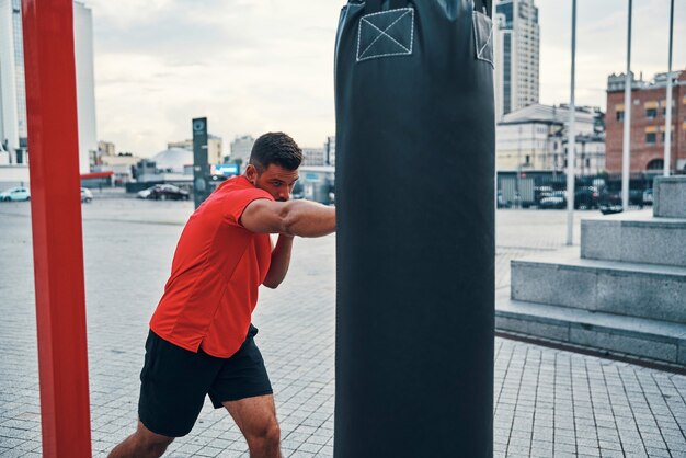 Foto forte giovane in abbigliamento sportivo che prende a calci il sacco da boxe mentre si esercita all'aperto