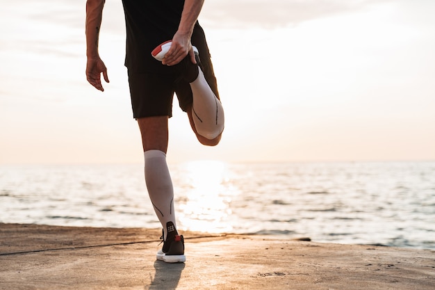 Strong young sportsman outdoors at the beach make stretching exercises.