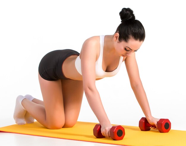 腕立て伏せをしている強い若い女性はダンベルで運動します。黄色いマットの上で激しいトレーニングをしているフィットネスモデル。