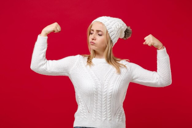 Foto forte giovane donna in maglione bianco, cappello isolato su sfondo muro rosso, ritratto in studio. stile di vita sano, emozioni della gente, concetto di stagione fredda. simula lo spazio della copia. mostra bicipiti, muscoli.
