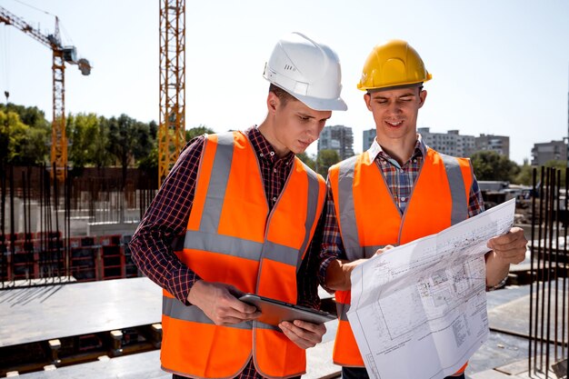 Structural engineer and construction manager dressed in orange work vests and helmets discuss documentation on the open air building site next to the crane .