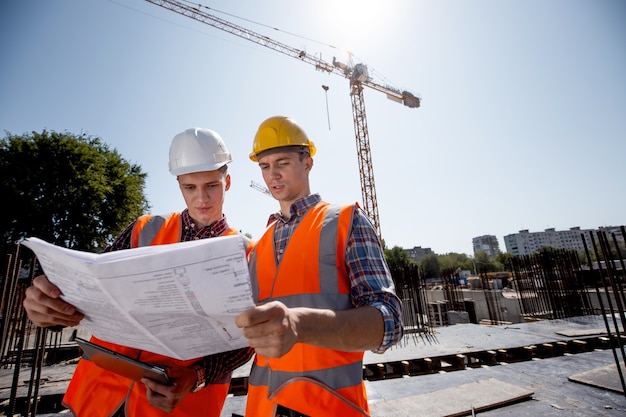 Structural engineer and construction manager dressed in orange work vests and helmets discuss documentation on the open air building site next to the crane .