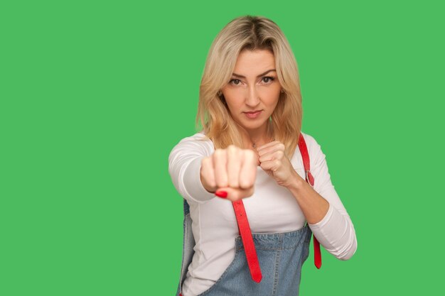 Struggle with threat Portrait of brave confident adult blond woman in stylish denim overalls boxing and fighting clenching fists to repel attack indoor studio shot isolated on green background