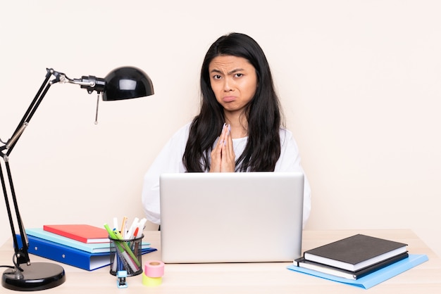 Student Aziatisch meisje op een werkplek met een laptop geïsoleerd op beige houdt palm bij elkaar. Persoon vraagt iets