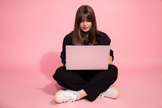 Student brown haired surprised charming Girl in casual clothes Holding Laptop Computer Sitting On Pink Background. Online Education Concept. Studio Shot. Looking at laptop.