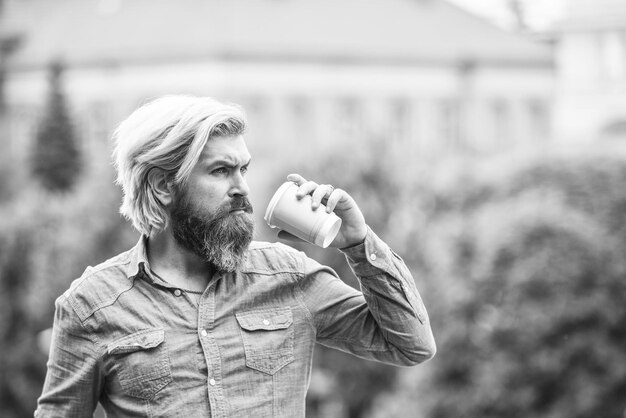 Student on coffee break bearded man drinking coffee while\
walking outdoors bearded hipster guy wearing casual clothes enjoys\
relaxing on weekend morning drinks concept