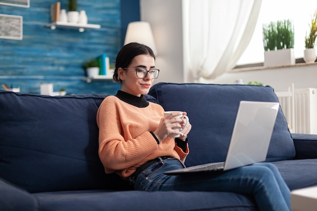 Student college learning management online les tijdens universitair seminar. Jonge vrouw die op de bank in de woonkamer zit en e-learningplatform controleert met behulp van een laptopcomputer