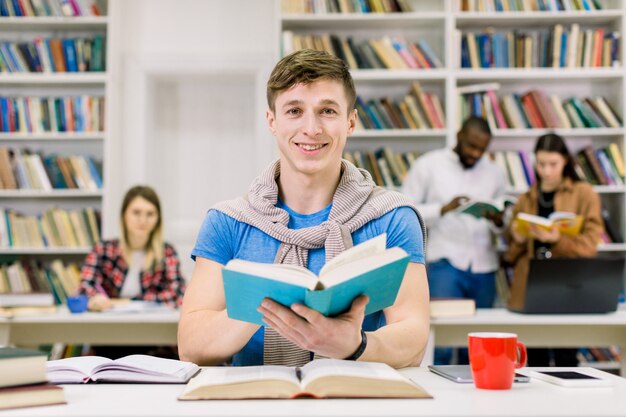 Student die camera bekijkt en glimlacht, geniet terwijl studerend in bibliotheek met zijn multi-etnische groepsgenoten op de ruimte