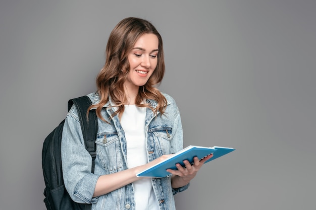 Student girl holding a notebook with homework in her hands reading and smiling isolated over grey wall background