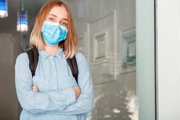 Student Girl met gekruiste armen op universiteitsinterieur tijdens coronavirus covid lockdow. Schoolmeisje in blauw masker met rugzak. Jonge vrouwelijke student met beschermend medisch masker.