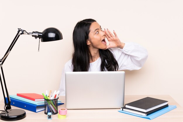 Ragazza dello studente in un posto di lavoro con un computer portatile isolato sul gridare beige con la bocca spalancata al laterale