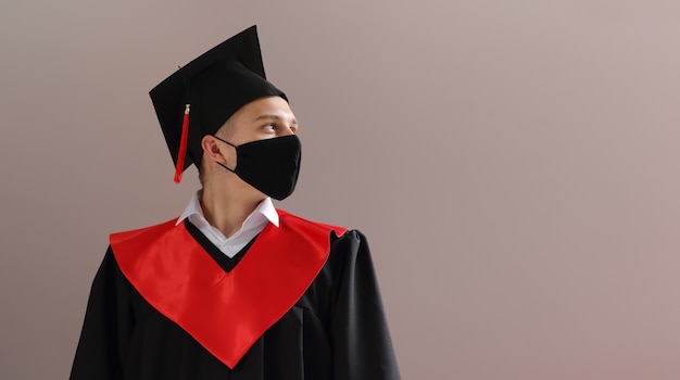 Photo a student in graduation gown and a square cap wear a medical face mask in a campus and look away