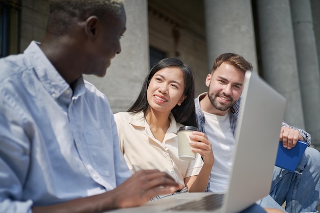 Photo student guy and his friends using a laptop while sitting on the steps