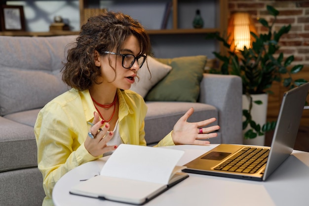 Student jong krullend meisje studeren thuis met behulp van webcam chat Videoconferentie bellen op laptopcomputer maken van aantekeningen praten op webcam Afstandsonderwijs concept