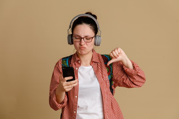 Student jonge vrouw in casual kleding dragen van een bril met koptelefoon en rugzak met mobiele telefoon kijken naar het scherm ontevreden met duim naar beneden staande over bruine achtergrond