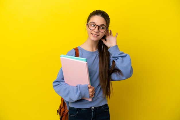 Student kind vrouw over geïsoleerde gele achtergrond luisteren naar iets door hand op het oor te leggen