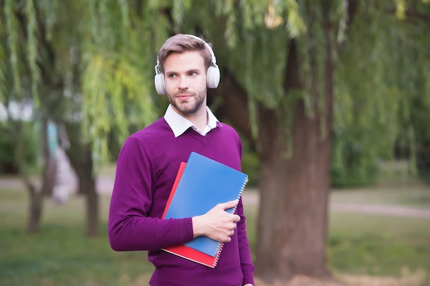 Student krijgt informatie uit boek. e-learning concept. man luistert naar muziek in een koptelefoon en houdt een notitieboekje vast. knappe kerel in oortelefoons ontspannen in het park. moderne levensopvoedingstechnologie.