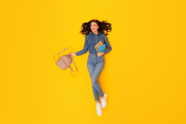 Student lady holding backpack and study materials over yellow background