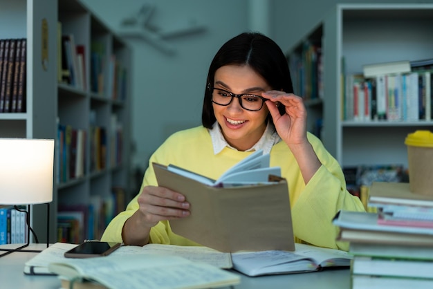 Student in library Brunette cheerful woman correcting her eyeglasses and reading book while sitting at the table during the evening with bookshelf at the background