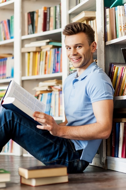 Student in library. Side view of handsome young man holding a book and smiling at camera while sitting on the floor and leaning at the library bookshelf