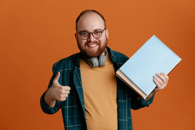 Photo student man in casual clothes wearing glasses with headphones holding notebooks looking at camera happy and positive smiling showing thumb up standing over orange background