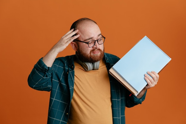 Student man in casual clothes wearing glasses with headphones holding notebooks looking at it confused holding hand on his head for mistake standing over orange background