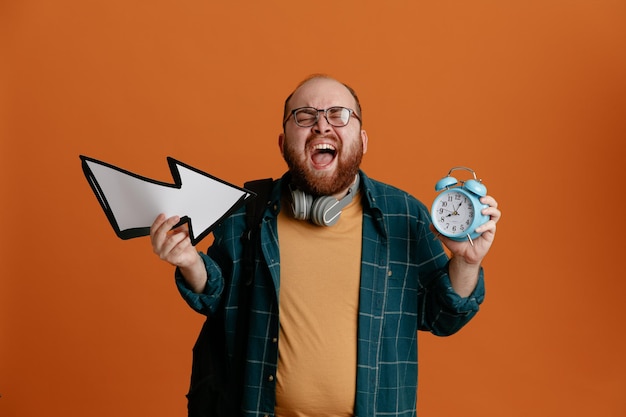 Foto student man in vrijetijdskleding met bril met koptelefoon met wekker en pijl gek, boos en gefrustreerd schreeuwen en schreeuwen staande over oranje achtergrond