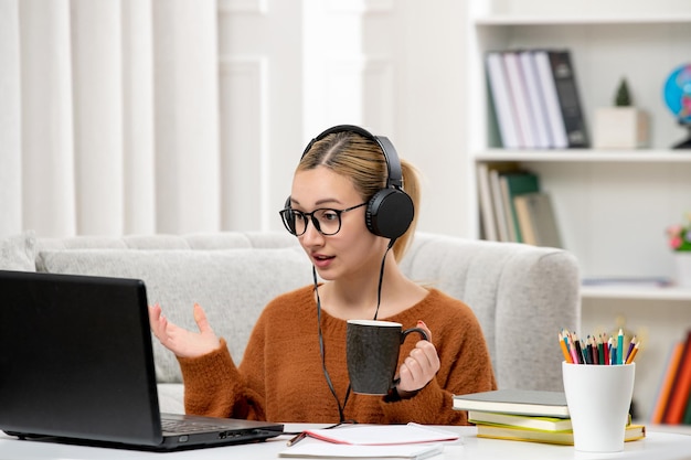 Foto studente online ragazza carina in occhiali e maglione che studia sul computer in possesso di una tazza di caffè