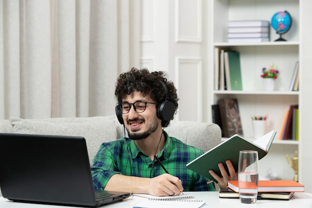 Student online cute young guy studying on computer in glasses in green shirt smiling with book