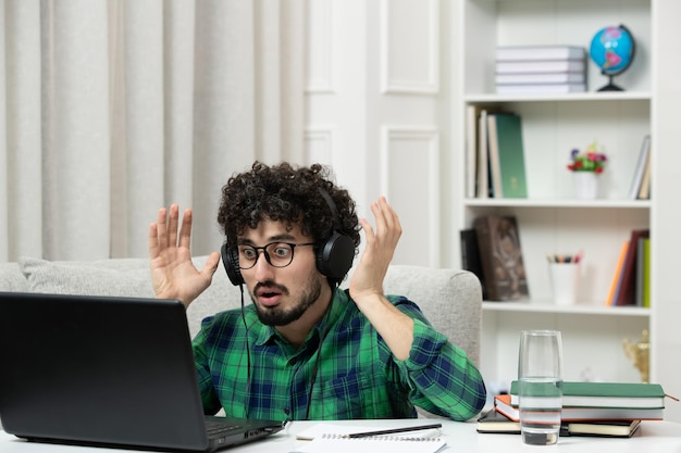 Student online cute young guy studying on computer in glasses in green shirt waving hands confusingly