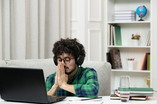 Student online schattige jonge kerel die op de computer studeert met een bril in een groen shirt die met gesloten ogen bidt