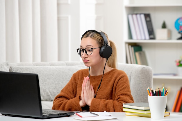 Studente online giovane ragazza carina con gli occhiali e maglione arancione che studia sul computer pregando