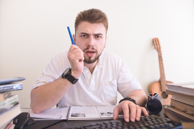 Student sitting at the table at home with a pen in his hands, looks at the computer monitor and writes.