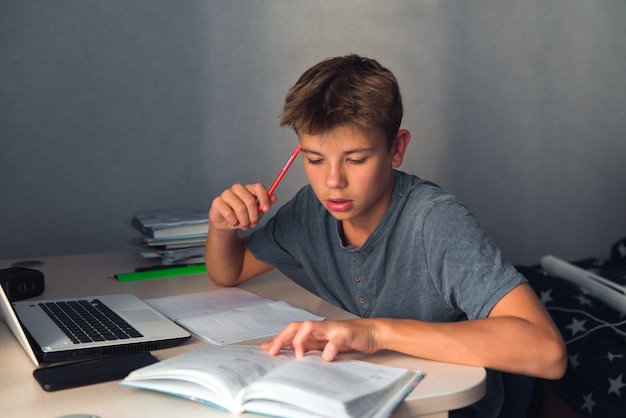 Ragazzo dell'adolescente dello studente che fa i compiti con il quaderno aperto del computer portatile e il posto di lavoro del computer a casa