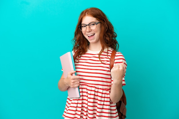 Student teenager redhead girl isolated on blue background celebrating a victory