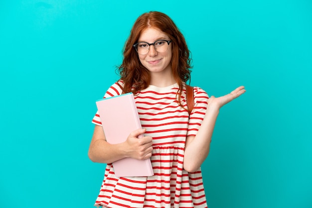 Student teenager redhead girl isolated on blue background having doubts while raising hands