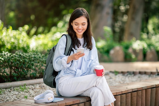 Student using smartphone while holding coffee in park