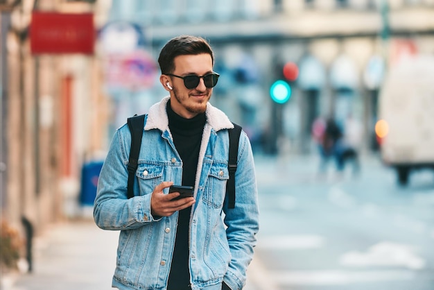 a student walking through the city carrying a backpack and wearing sunglasses while using a