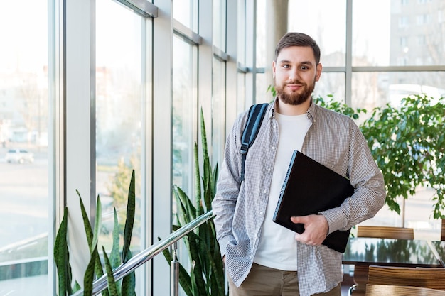 Student with backpack and laptop in reopen university campus near window. Caucasian teenager, confident bearded man carrying laptop in library. Freelancer in modern coworking office with plants.