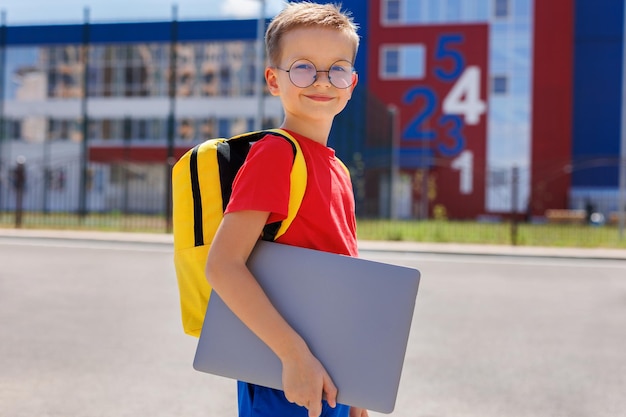 A student with glasses and a backpack holds a laptop in his hands against the background of the school