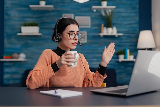 Student with glasses casually waving hello in laptop videocall. Person with headphones raising hand in remote video call lecture. College girl attendig online class from home office.