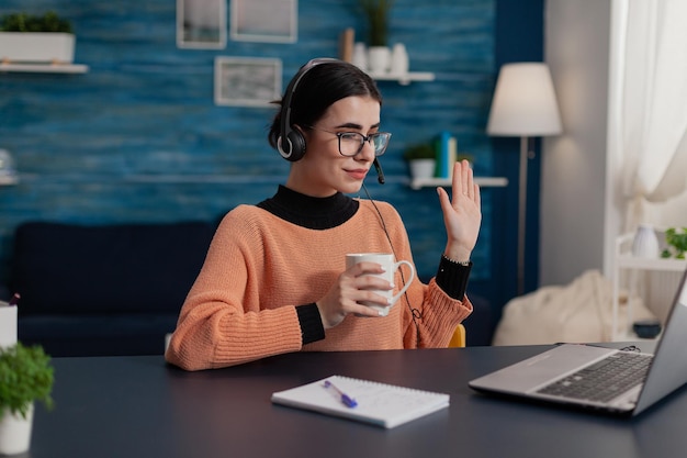 Student with headphones waving hello in laptop videocall. College girl with glasses holding cup attendig online video call lecture. Programmer raising hand in remote teleconference course.