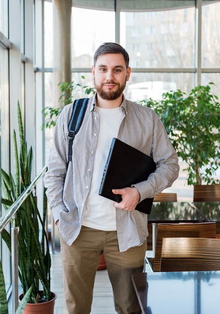 Studente con laptop e zaino vicino alla finestra nel campus universitario riaprire. adolescente caucasico, uomo barbuto sicuro che trasporta laptop in biblioteca. libero professionista in un moderno ufficio di coworking con piante.