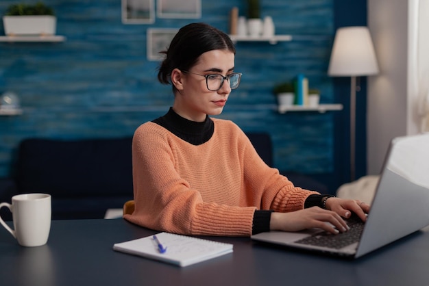 Student with notebook studying remote looking at laptop screen. Influencer with glasses typing on keyboard sitting at home desk. College girl browsing the internet. Freelancer sending email.