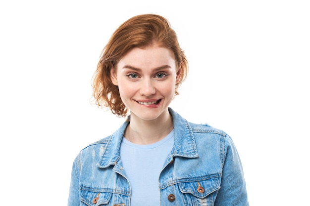 A student with red hair thinks happily, bites her lip and looks straight. Pensive woman in a blue denim jacket on a white background. Copy space