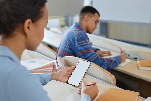 Student writing off from the smartphone sitting at table with books during exam in university